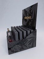 A black display stand filled with rectangular black cosmetic tubes, with a few small bottles on the left side. The stand has a tropical leaf pattern and the word 'SABOTAGE' in gold lettering on the back panel, photographed on a light gray studio background.