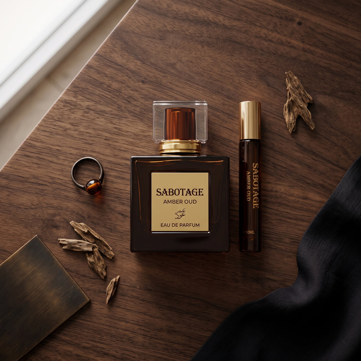 Sabotage Amber Oud Eau de Parfum bottle with matching travel spray, tiger's eye ring, agarwood chips, and black silk fabric on a rustic wooden table.